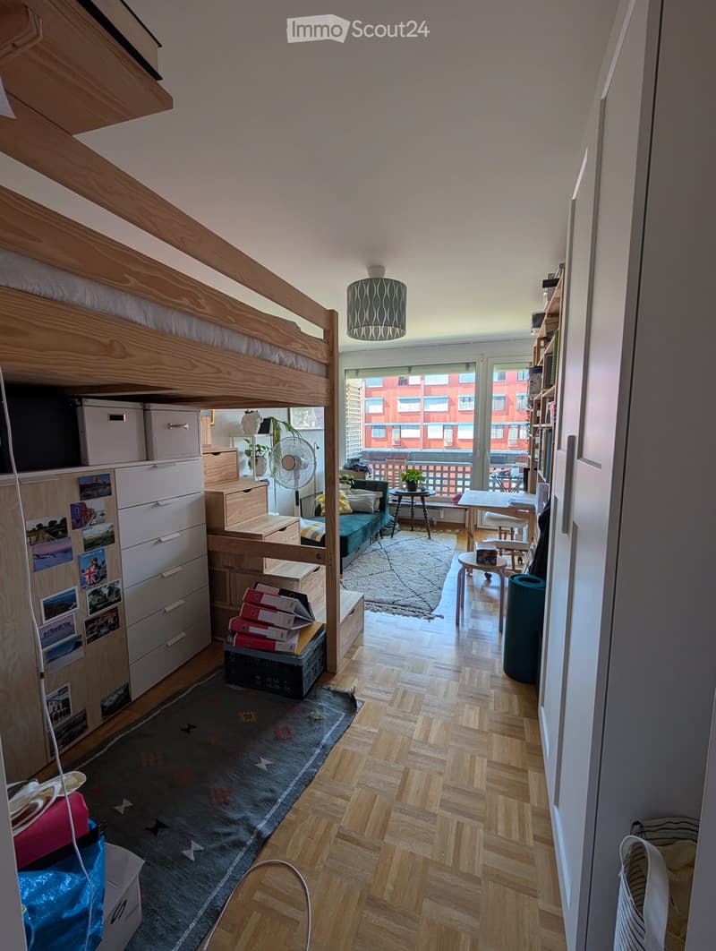A furnished room with a bunk bed, sofa, small table, and various storage items. The room features wooden flooring, a large window with a view, and a balcony area with outdoor furniture.