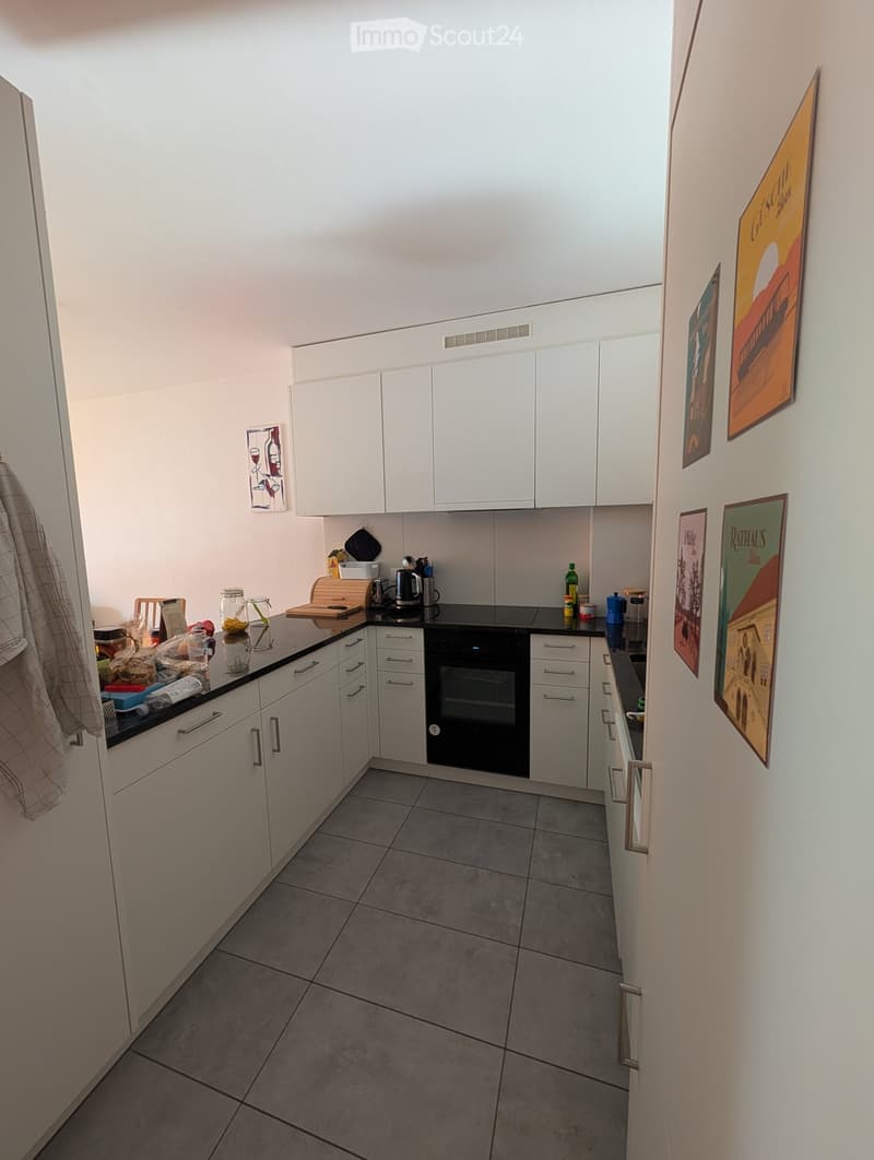 Modern kitchen with white cabinets, black countertop, and various appliances including an oven and a refrigerator. The space is well-lit with a tiled floor and several decorative posters on the wall.