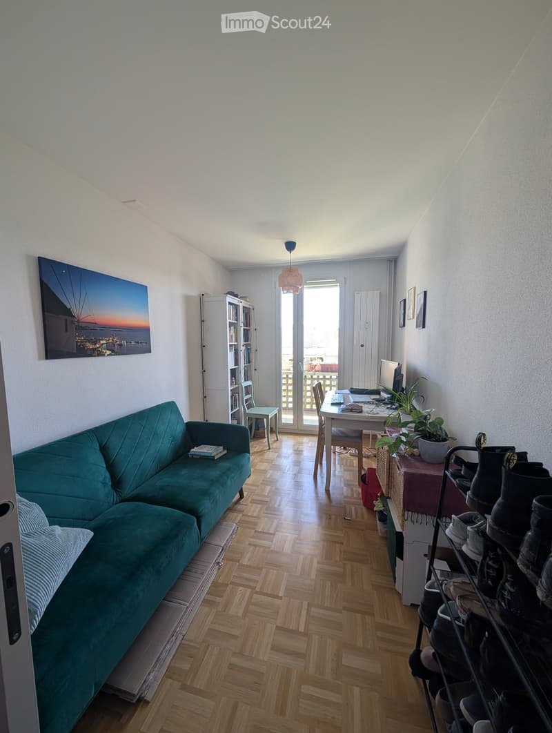 A modern living room with a teal sofa, wooden floor, a small table with a laptop, a bookshelf, and a shoe rack near the entrance.