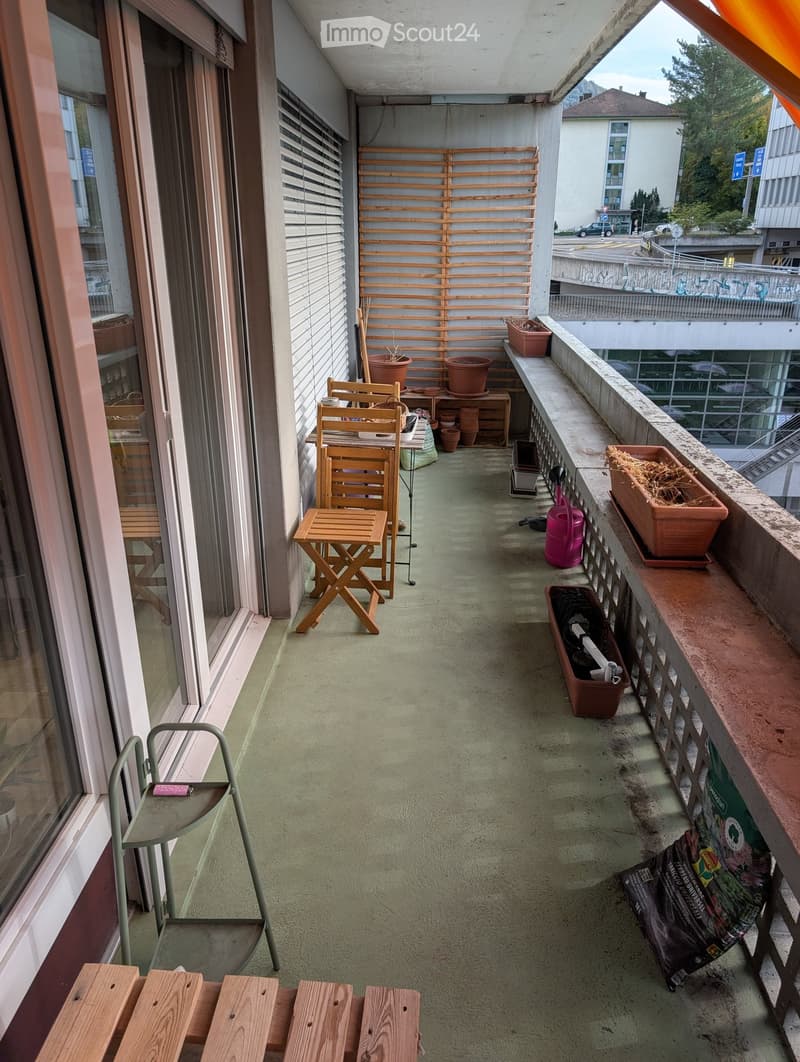 Balcony with wooden table, chairs, and several potted plants, with a view of the street and surrounding buildings.