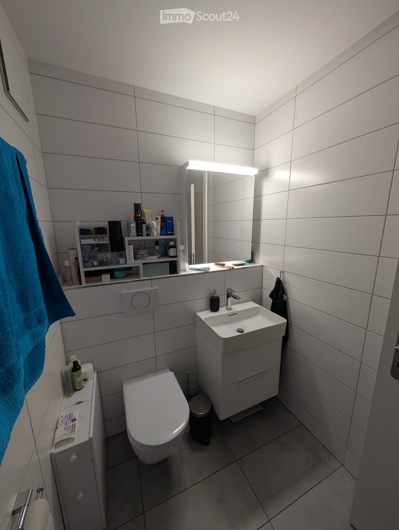 Modern bathroom with white tiles, a sink with a modern faucet, a toilet, a large mirror with built-in lighting, shelves with toiletries, and a towel hanging on the wall.