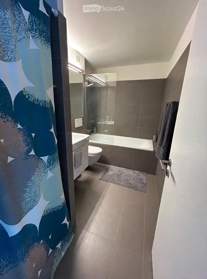Modern bathroom with a bathtub, toilet, sink, and mirror. Features tiled walls, a shower curtain, and a small rug on the floor.