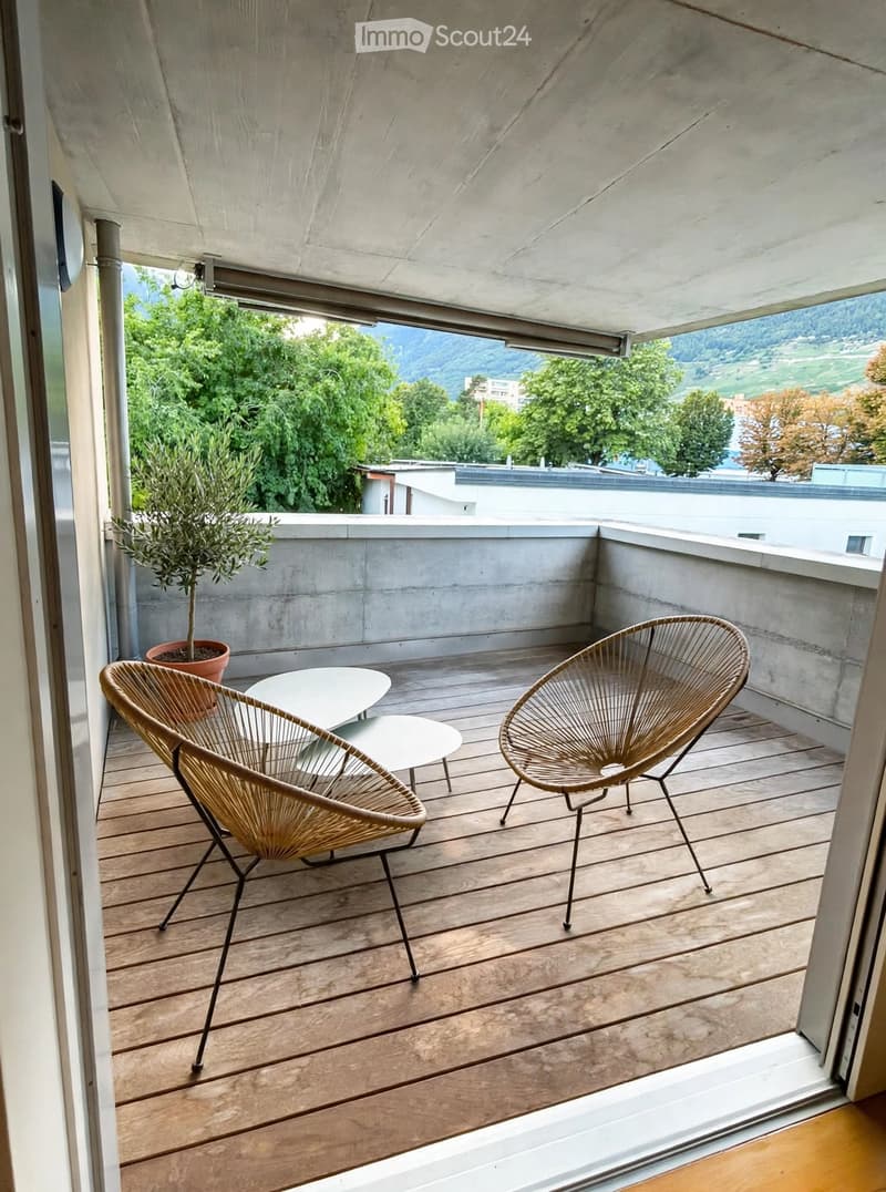 Wooden deck with two wicker chairs, two small white tables, and a potted plant, overlooking a scenic view of trees and hills.