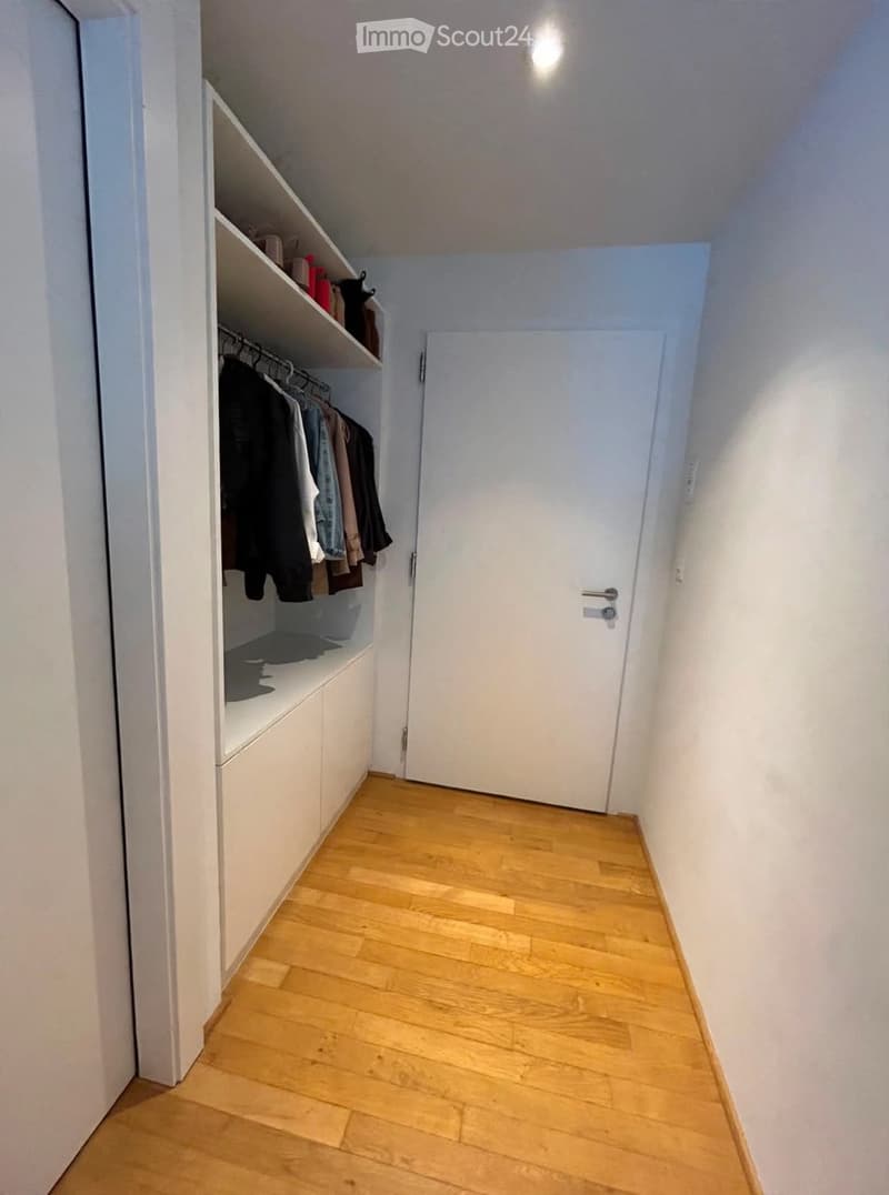 A narrow hallway with wooden flooring, white walls, and a built-in closet with hanging clothes and shelves. A closed white door is visible at the end of the hallway.