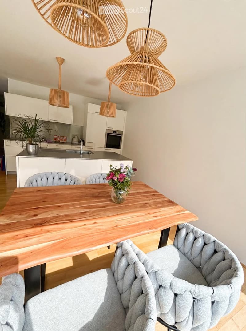 Modern dining area with a wooden dining table, grey upholstered chairs, a vase with flowers, and a sleek kitchen in the background featuring white cabinets, an oven, and a sink.