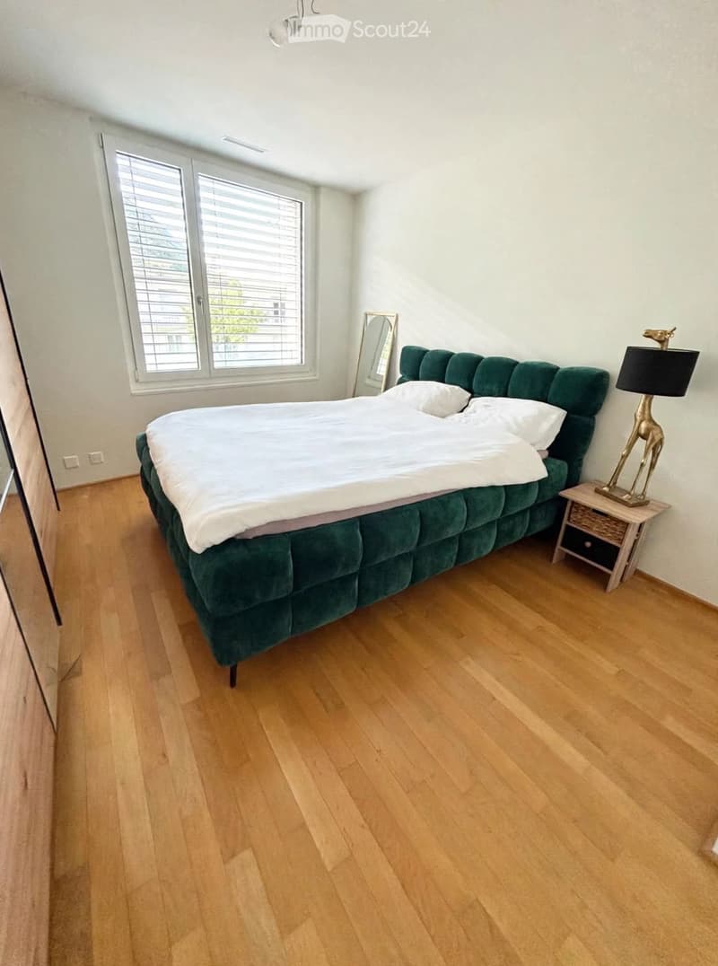 Bright bedroom with wooden flooring, large window with blinds, green upholstered bed, nightstand with lamp, and decorative mirror.