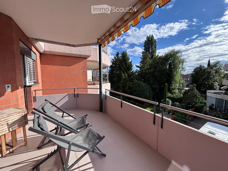 Balcony with wooden table, two folding chairs, metal railing, and awning, overlooking a tree-lined area.