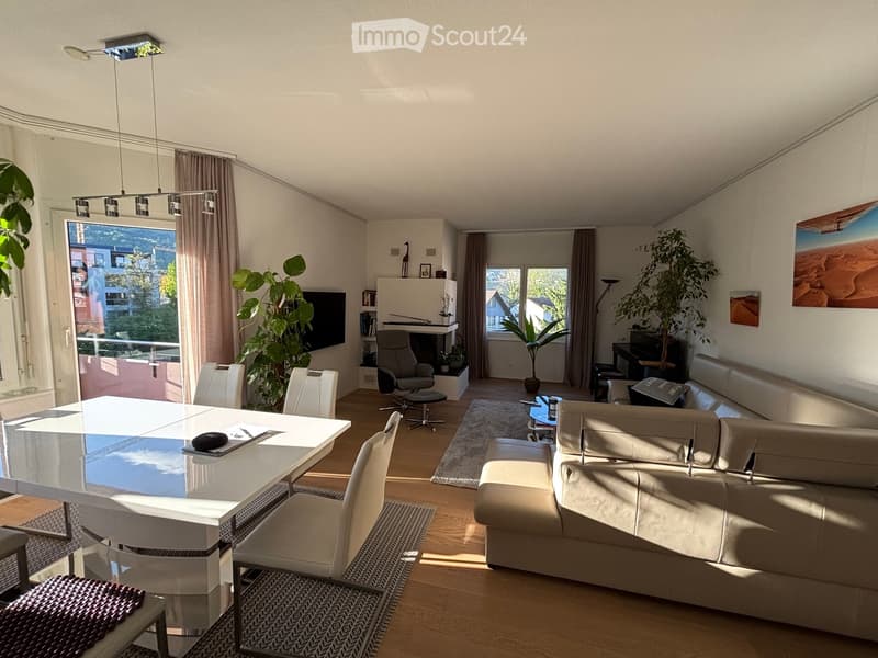 Modern living room with a large window offering a scenic view, featuring a cream-colored sofa, a glass dining table with white chairs, a fireplace, and decorative plants.