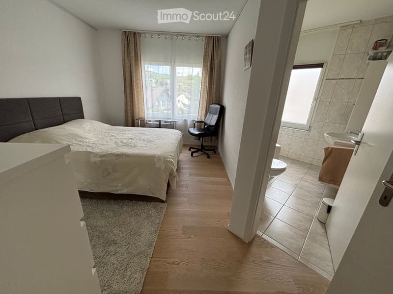 Bedroom with a large bed, a chair, a small table, and an adjacent bathroom with a sink and toilet.