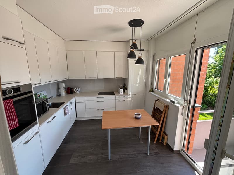 Modern kitchen with white cabinets, a wooden dining table, and various appliances including an oven, microwave, and fridge. The space features large windows and a view of greenery outside.