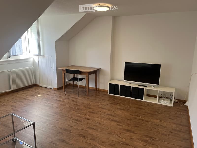 Bright, slanted-ceiling room with wooden flooring, a TV on a white stand, a small wooden desk with a chair, and a glass coffee table.