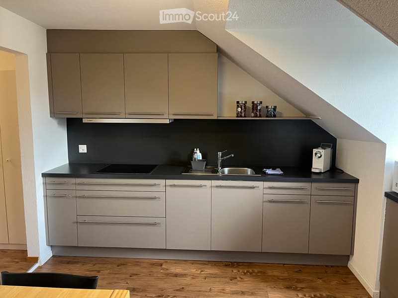 Modern kitchen with light grey cabinets, black countertops, stainless steel sink, and various appliances. Wooden floor and a small dining area visible.