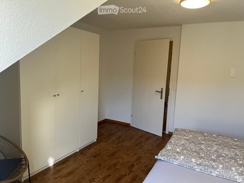 A small, furnished room with a wooden floor, a bed with a patterned bedspread, a white wardrobe, and a wicker chair.
