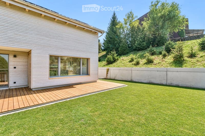 A modern house with a raised wooden deck, large window, and well-maintained lawn surrounded by trees and a concrete wall.