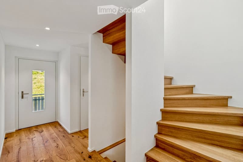 A modern hallway with wooden flooring, white walls, recessed lighting, and a staircase with wooden steps and railings. Two doors are visible, one with a small window showing greenery outside.