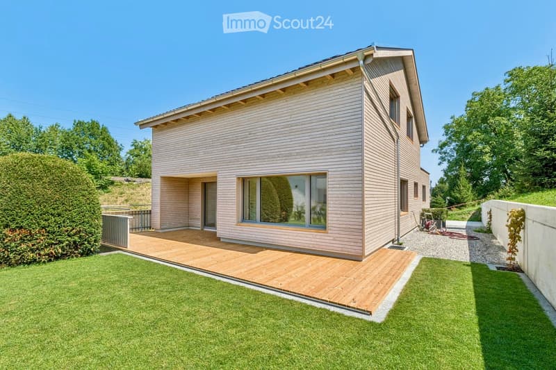 Modern single-story house with light wood siding, large windows, and a wooden deck extending from the entrance. The surrounding area features a well-maintained lawn and a raised wooden deck.