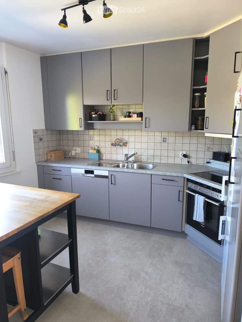 Modern kitchen with grey cabinets, a central island, a sink, and an oven. The space includes a wooden table with a stool, a refrigerator, and a dishwasher.