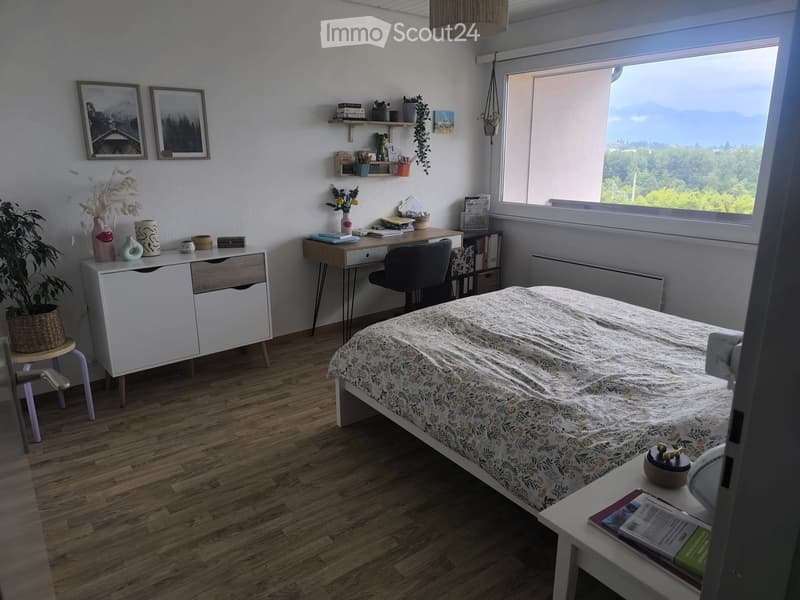 Cozy bedroom with a double bed, nightstand, desk, and cabinet. Features a large window with a scenic view, plants, and decorative items.