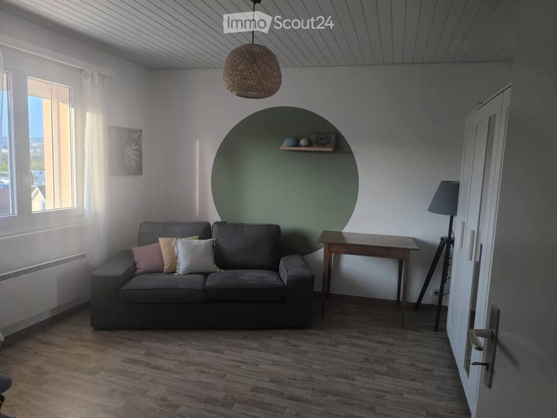 Modern living room with wooden flooring, grey sofa with throw pillows, small wooden table, floor lamp, and a decorative shelf on a green accent wall.