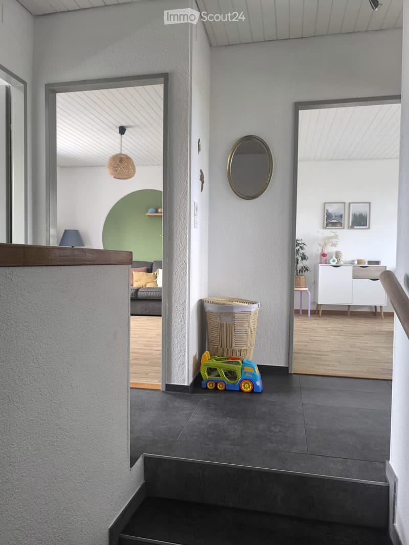 Modern hallway with grey tiled floor, white walls, and a staircase leading upwards. Features include a round mirror, a woven basket, a small toy car, and a view into a living area with a sofa and green accent wall.