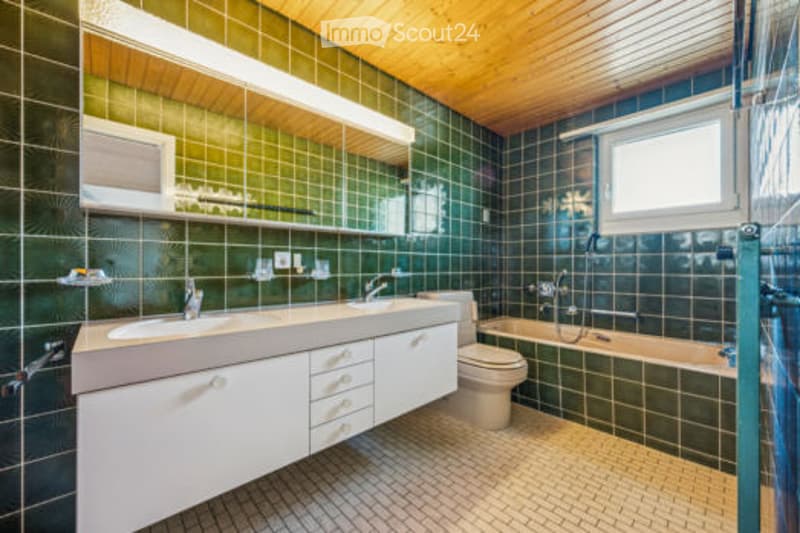 Modern bathroom with green tiled walls, white cabinetry, double sink, bathtub, and a large window for natural light.