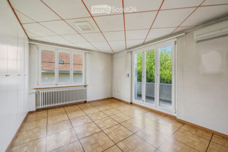 Empty room with wooden flooring, white walls, large window with view, sliding glass door leading to balcony, radiator, and air conditioning unit.