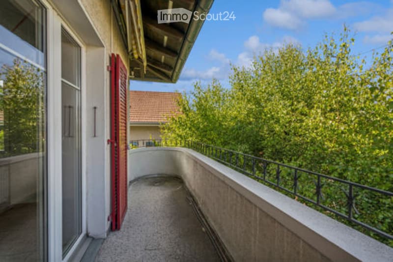 Balcony with a view of green trees, featuring a concrete floor, metal railing, and a red shuttered door leading to the interior.