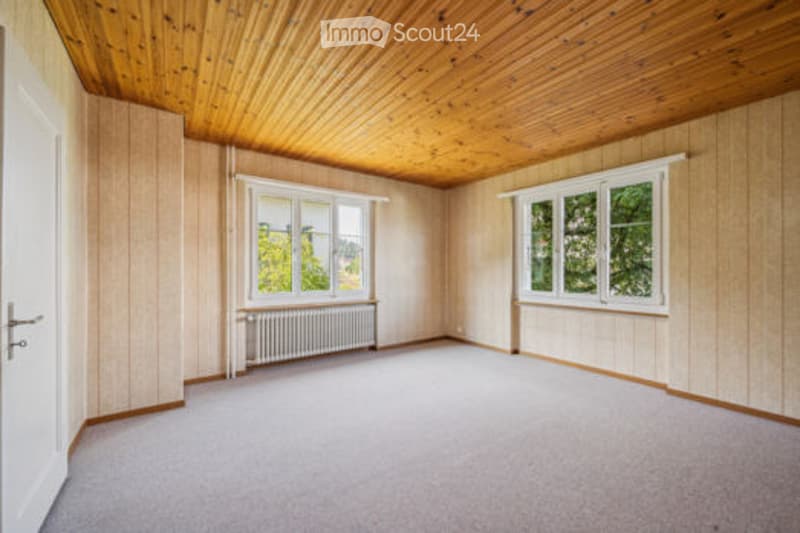 Empty room with wooden panel walls and ceiling, large windows, radiator, and carpeted floor.