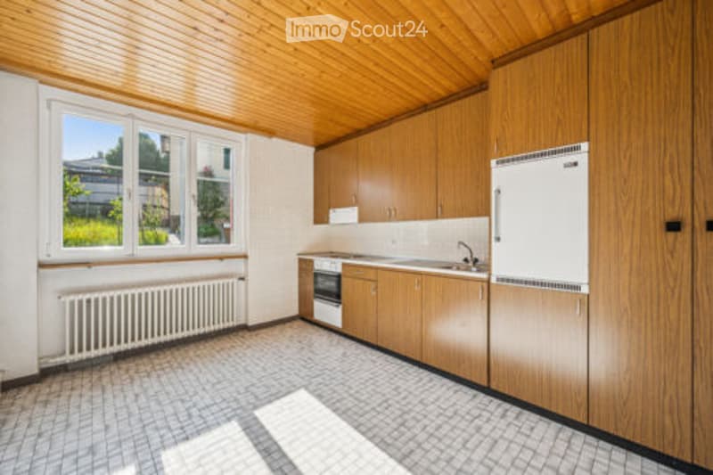 Empty kitchen with wooden cabinets, tiled floor, and large windows. No appliances or furniture present.