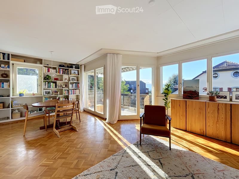 Bright living room with wooden flooring, large windows, a round wooden dining table with chairs, a bookshelf, a cabinet, and a comfortable armchair.