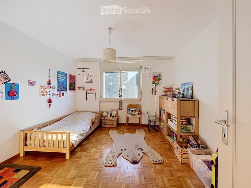 A cozy children's bedroom with a wooden bed, bookcase, small chair, toy cart, and decorative rug. The room features wooden flooring, white walls, and large windows with a view of trees.
