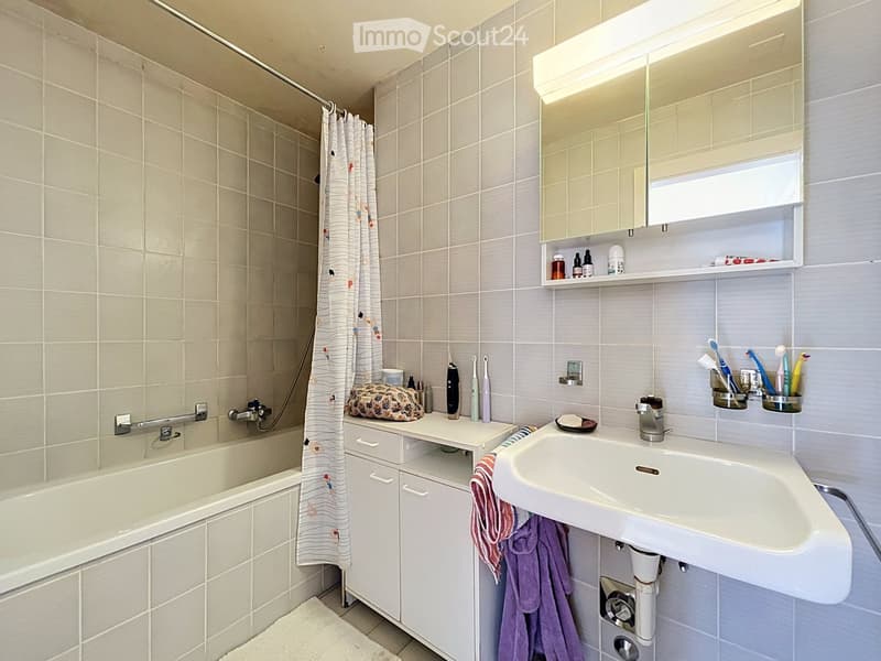 Modern bathroom with a bathtub, sink, mirror, and cabinet. Includes a shower curtain, towels, and personal care items on the sink counter.