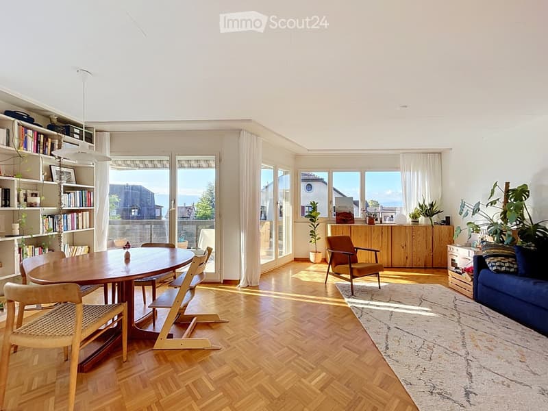 Bright, open living and dining area with wooden floors, large windows, and a view. Features a blue sofa, wooden dining table with chairs, bookshelf, and potted plants.