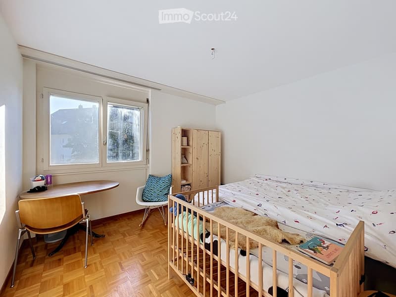 A cozy bedroom with a wooden floor, a double bed with a colorful blanket, a wooden wardrobe, a small table with a chair, and a baby cradle with a stuffed animal.