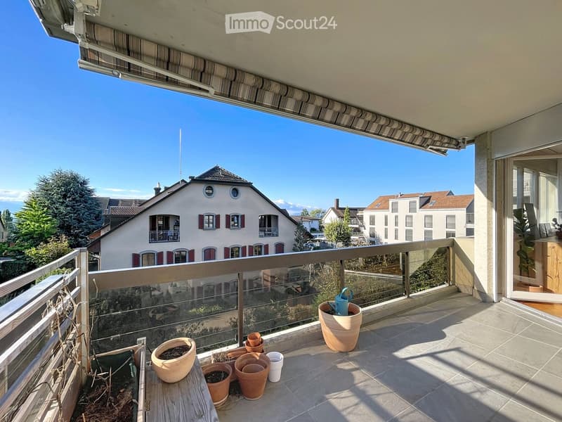 Balcony with tiled floor, glass railing, and several potted plants, including a watering can.