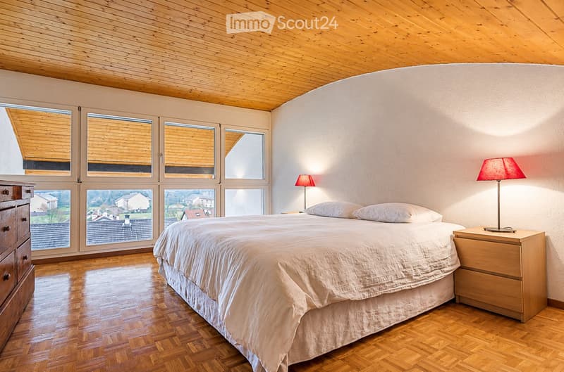 Bright bedroom with wooden flooring, large windows, a neatly made bed, nightstands with lamps, and a wooden dresser.