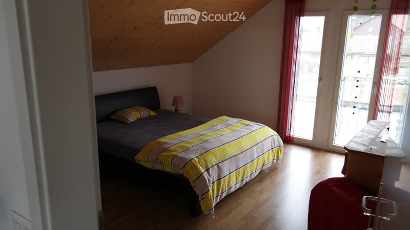Cozy attic bedroom with wooden floors, slanted ceiling, large window, and sliding glass door leading to a balcony.