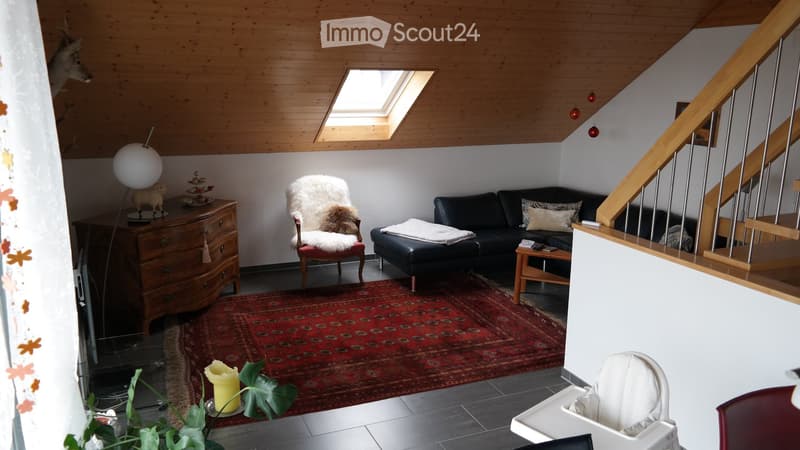 A cozy living room with wooden beams, a skylight, a red patterned rug, a black leather sofa, a white fur chair, a wooden side table, a floor lamp, and a staircase leading upwards.
