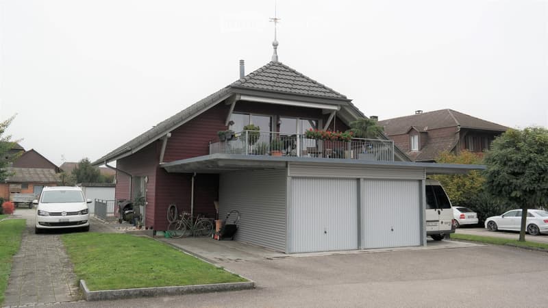 Two-story house with red siding, grey garage, balcony with plants, and parked vehicles.