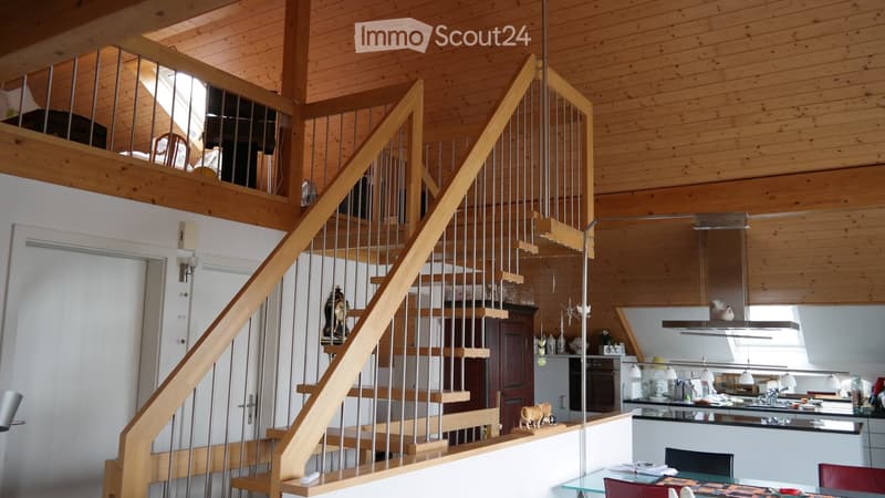 Open-plan living space with a wooden staircase leading to an upper level, a modern kitchen area with stainless steel appliances, and a dining area with a glass table and chairs.