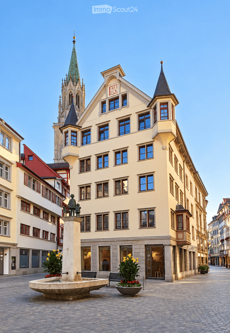 A multi-story building with a light yellow facade, multiple windows, and ornate architectural details. The building has a sign reading "FREI ECK" on the roof. A small fountain with a statue and potted plants is in front of the building, and a church spire