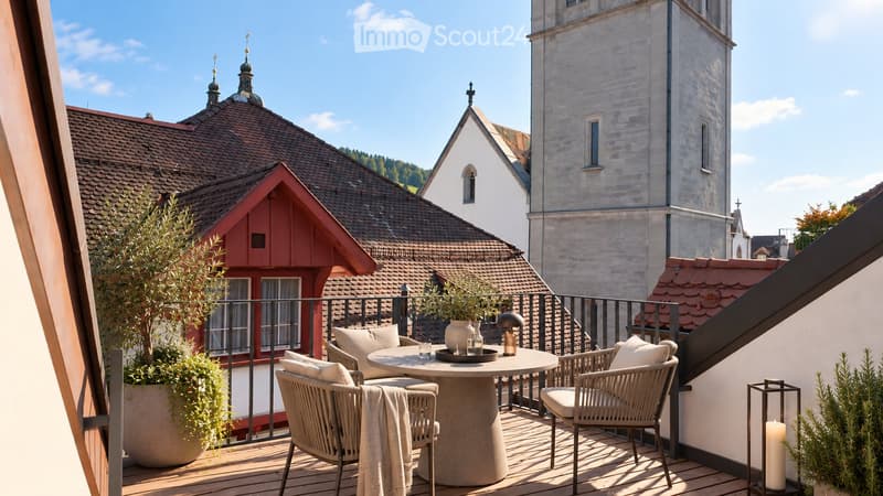 Balcony with wooden decking, round table, four chairs, cushions, vase, candle, and potted plants, overlooking a historic town with a church tower.