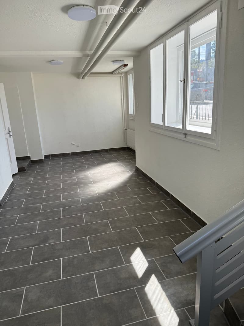 Empty hallway with grey tiled floor, white walls, and large windows allowing natural light. Features a staircase railing on the right and a door on the left.