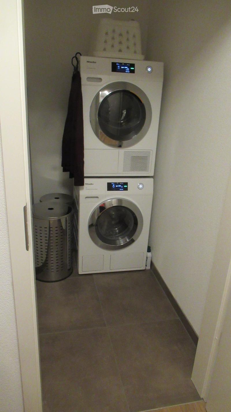 A small room with a white washing machine and a white dishwasher stacked on top of each other, a metal trash bin, and a white laundry basket on top of the washing machine.