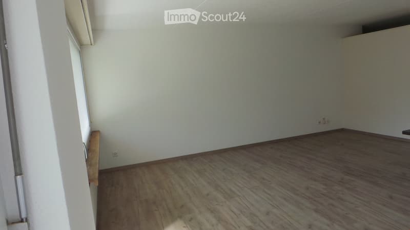 Empty room with wooden flooring and white walls, featuring a small wooden bench and a door frame visible on the left side.