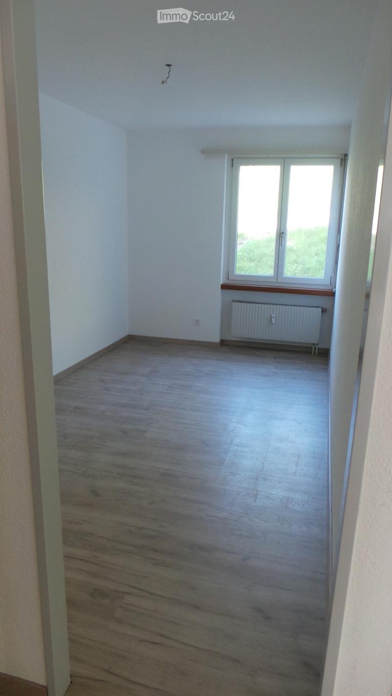 Empty room with wooden flooring, white walls, a window with white frame, and a radiator below the window.