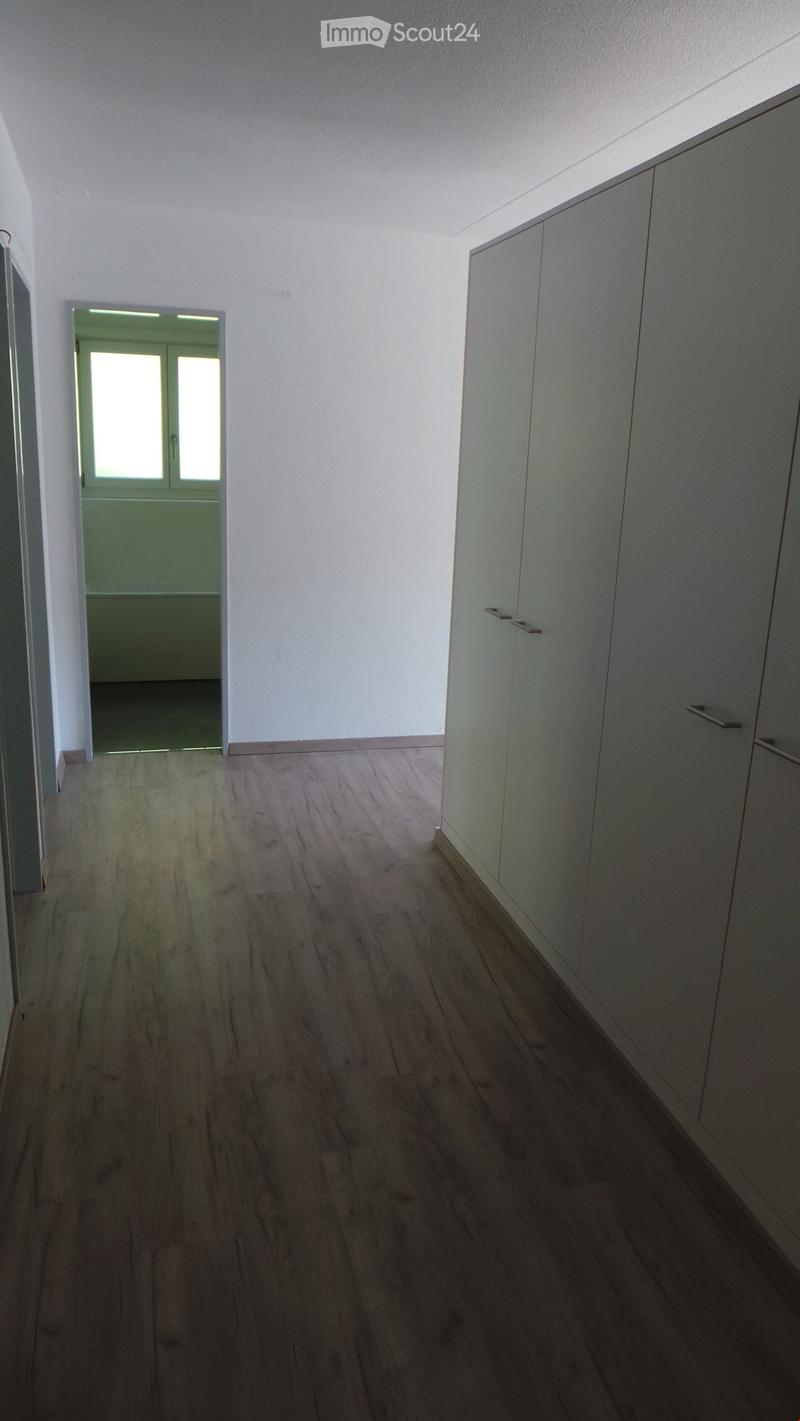 Empty hallway with wooden flooring, white walls, and built-in cabinets on one side. A doorway leads to another room with green walls and a window.