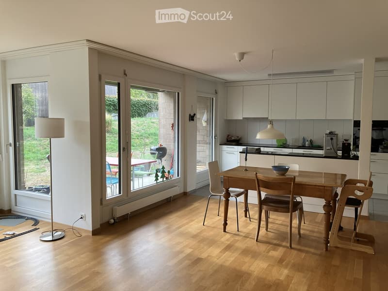 Bright, open-plan living and dining area with wooden flooring, large windows, a dining table with chairs, a floor lamp, and a modern kitchen area with cabinets and a pendant light.