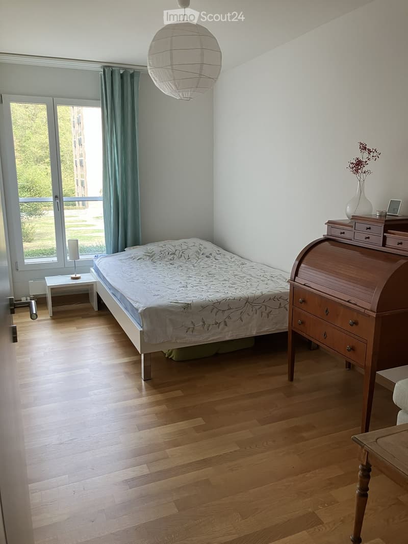 Bright bedroom with wooden flooring, a double bed with a patterned duvet, a nightstand with a lamp, a wooden dresser with drawers, and a vase with dried flowers on top.