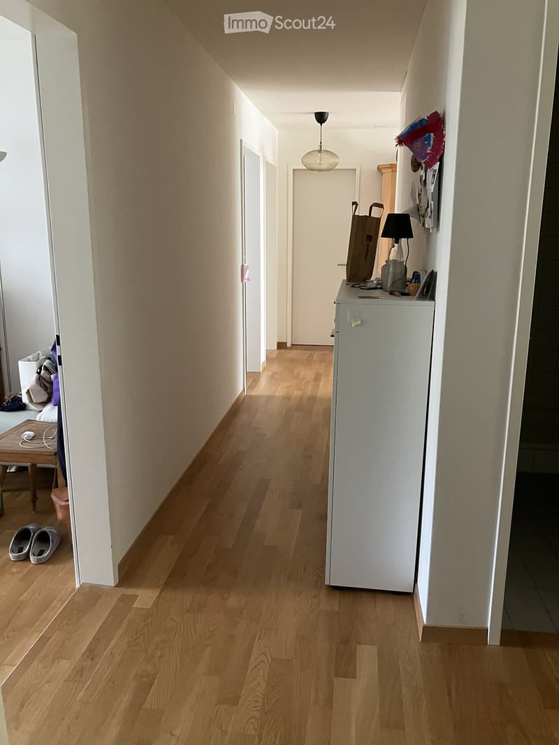 A narrow hallway with wooden flooring, white walls, and a hanging light fixture. A small table with a lamp and a shopping bag is visible on the right side.
